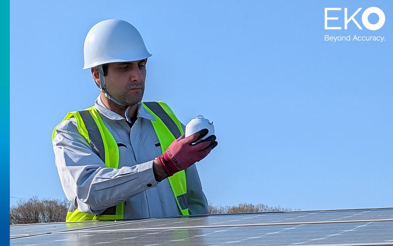 A technician wearing a hard hat, safety vest, and gloves carefully inspects an EKO MS-80SH pyranometer outdoors next to a solar panel array under a clear blue sky. The EKO 'Beyond Accuracy' logo is in the top right corner.